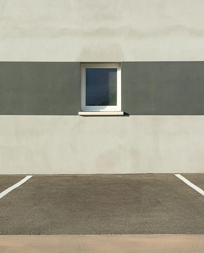 A modern building facade with three windows, featuring a minimalist design and an empty parking lot in the foreground.