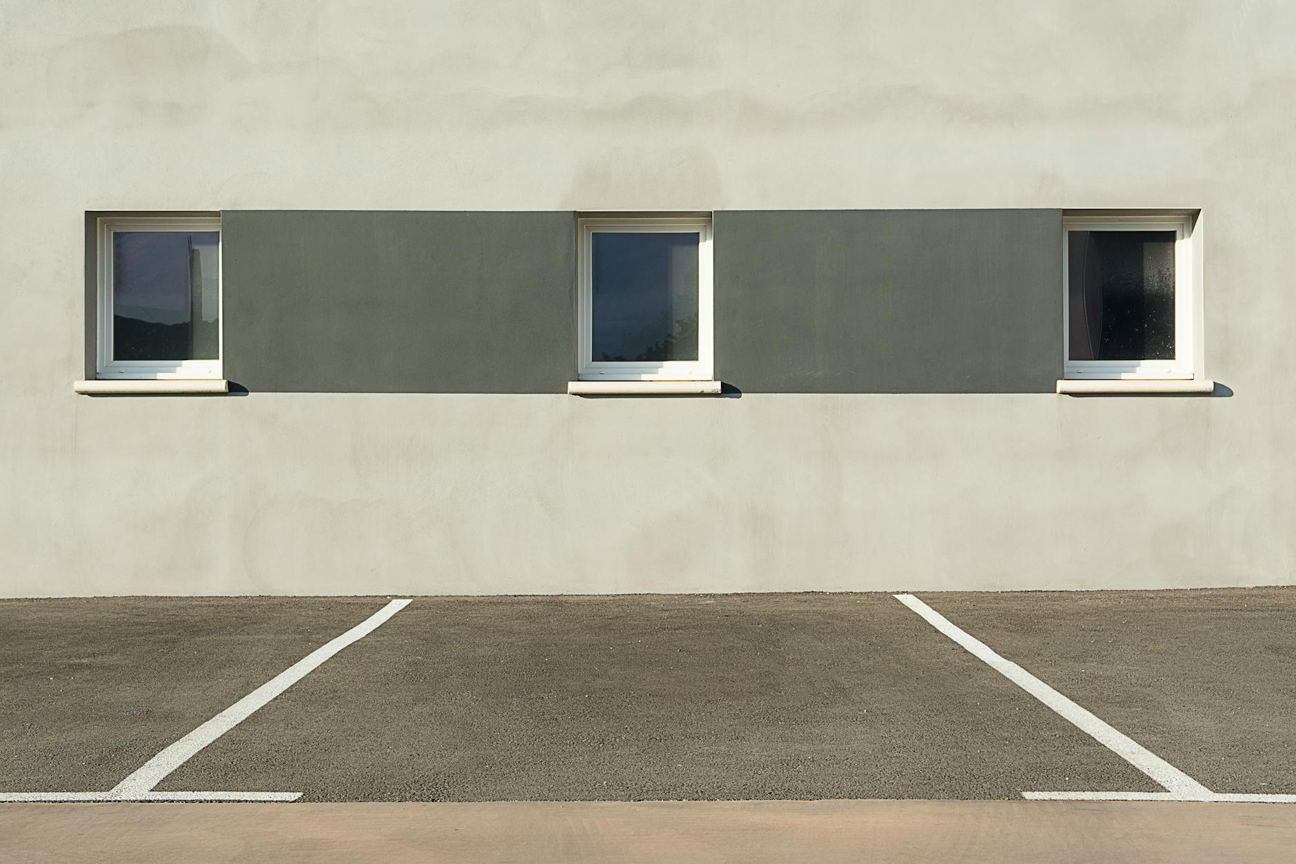 A modern building facade with three windows, featuring a minimalist design and an empty parking lot in the foreground.