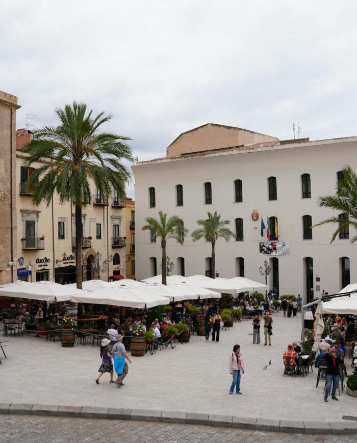 Busy Piazza Del Duomo in Cefalù, Italy, bustling with people and cafes under a cloudy sky.