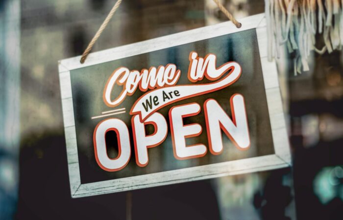 A welcoming 'Come in, We Are Open' sign hanging on a glass window of an urban shop.