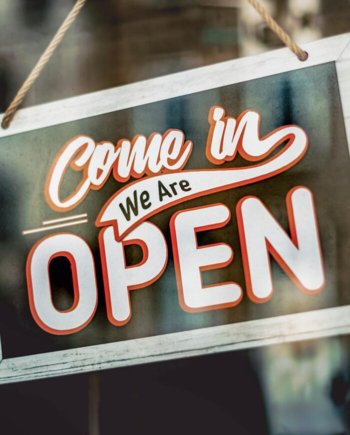 A welcoming 'Come in, We Are Open' sign hanging on a glass window of an urban shop.