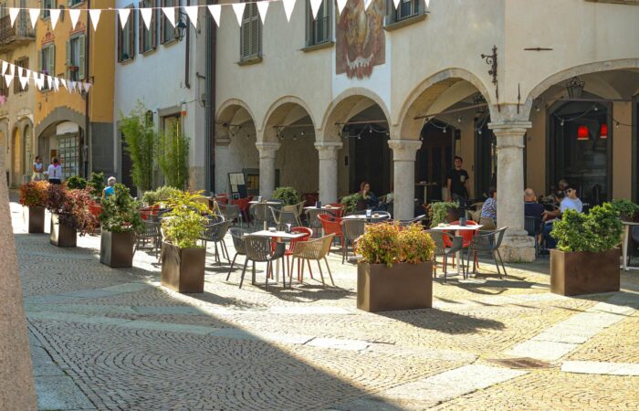 A picturesque outdoor café with vibrant seating and plants in a sunlit European piazza. A picturesque outdoor café with vibrant seating and plants in a sunlit European piazza.