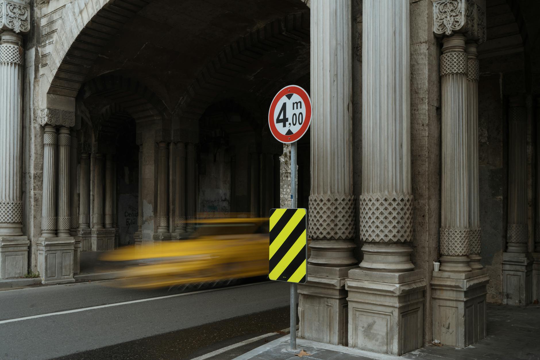 A yellow taxi speeds through a stone tunnel with classic columns and arches, creating a blur of motion.