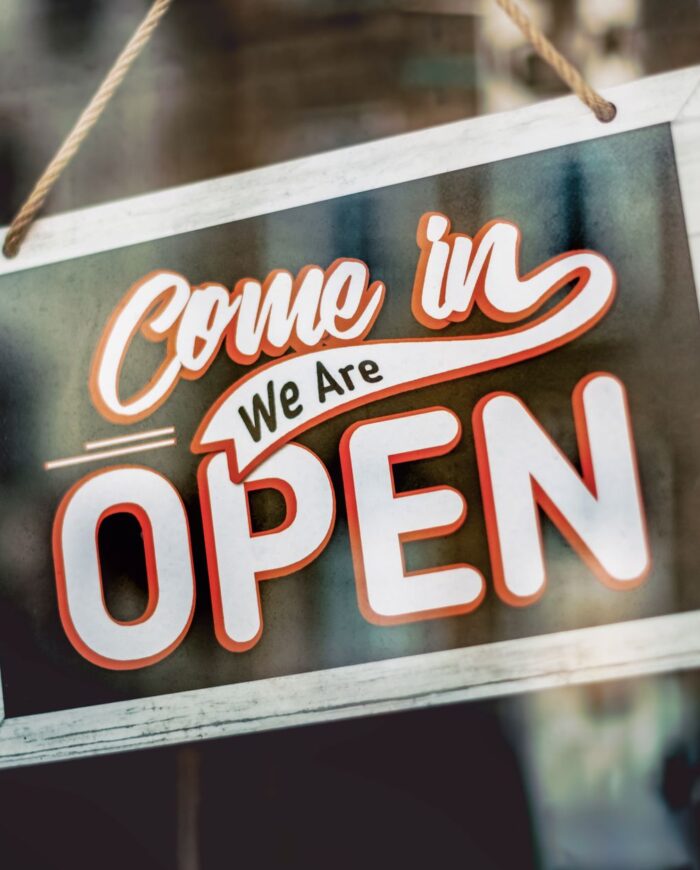 A welcoming 'Come in, We Are Open' sign hanging on a glass window of an urban shop.