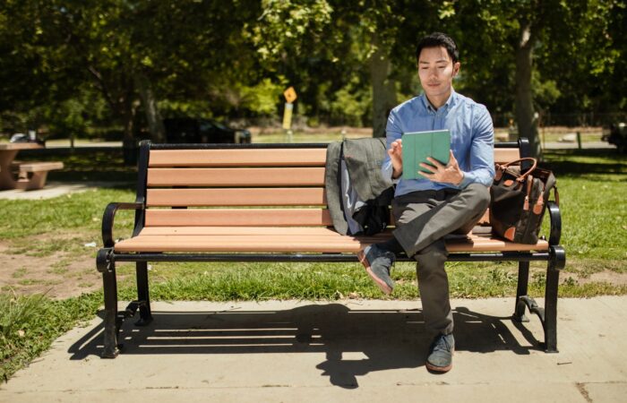 An adult man sits on a bench in a park, using a tablet on a sunny day.