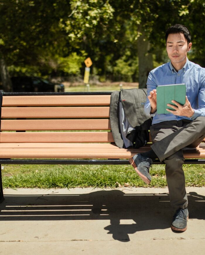 An adult man sits on a bench in a park, using a tablet on a sunny day.