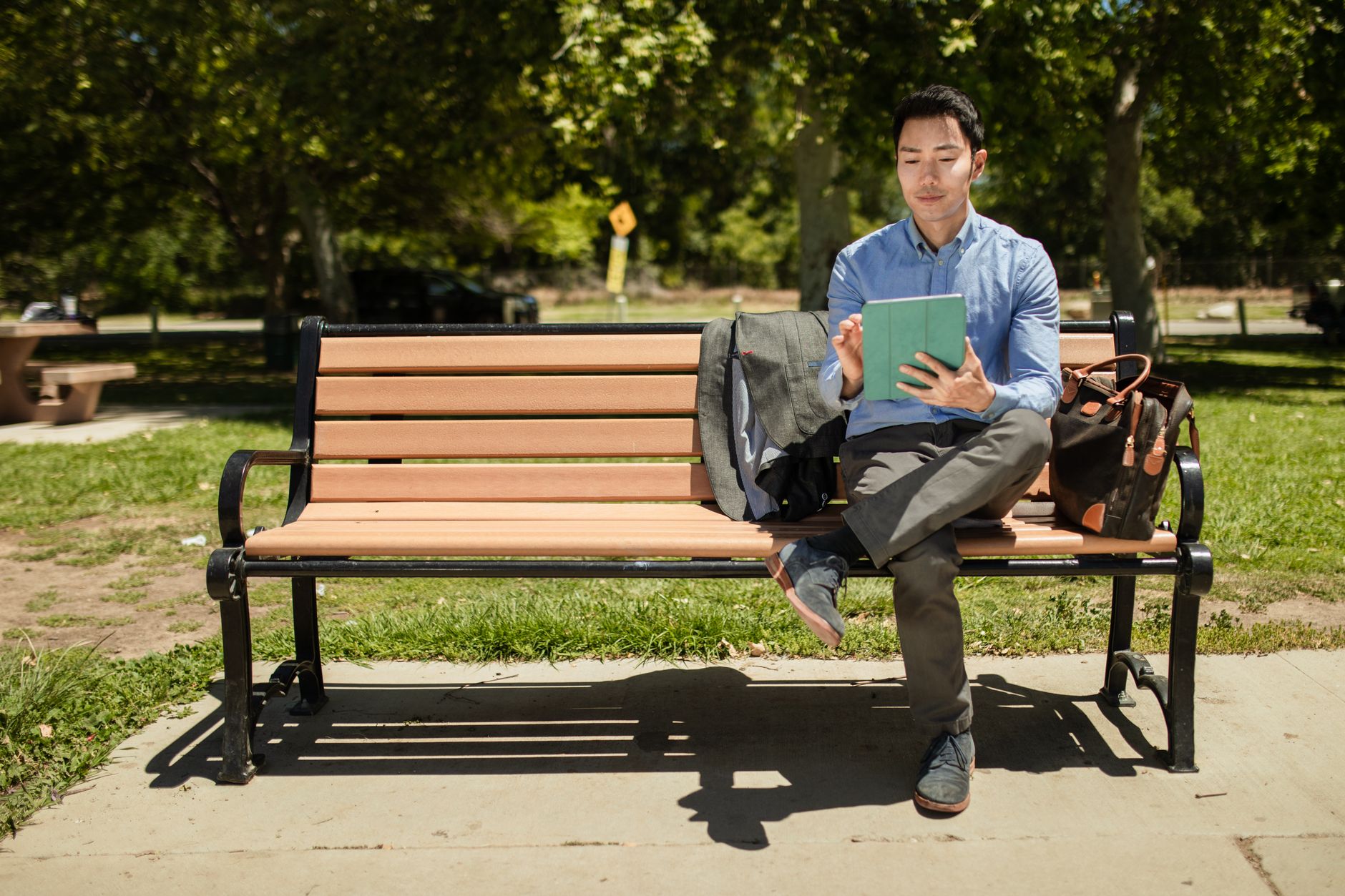 An adult man sits on a bench in a park, using a tablet on a sunny day.