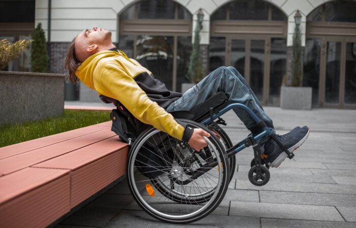 A young man in a hoodie relaxes in a wheelchair outside a modern building. A young man in a hoodie relaxes in a wheelchair outside a modern building.