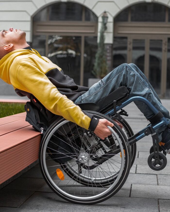 A young man in a hoodie relaxes in a wheelchair outside a modern building.